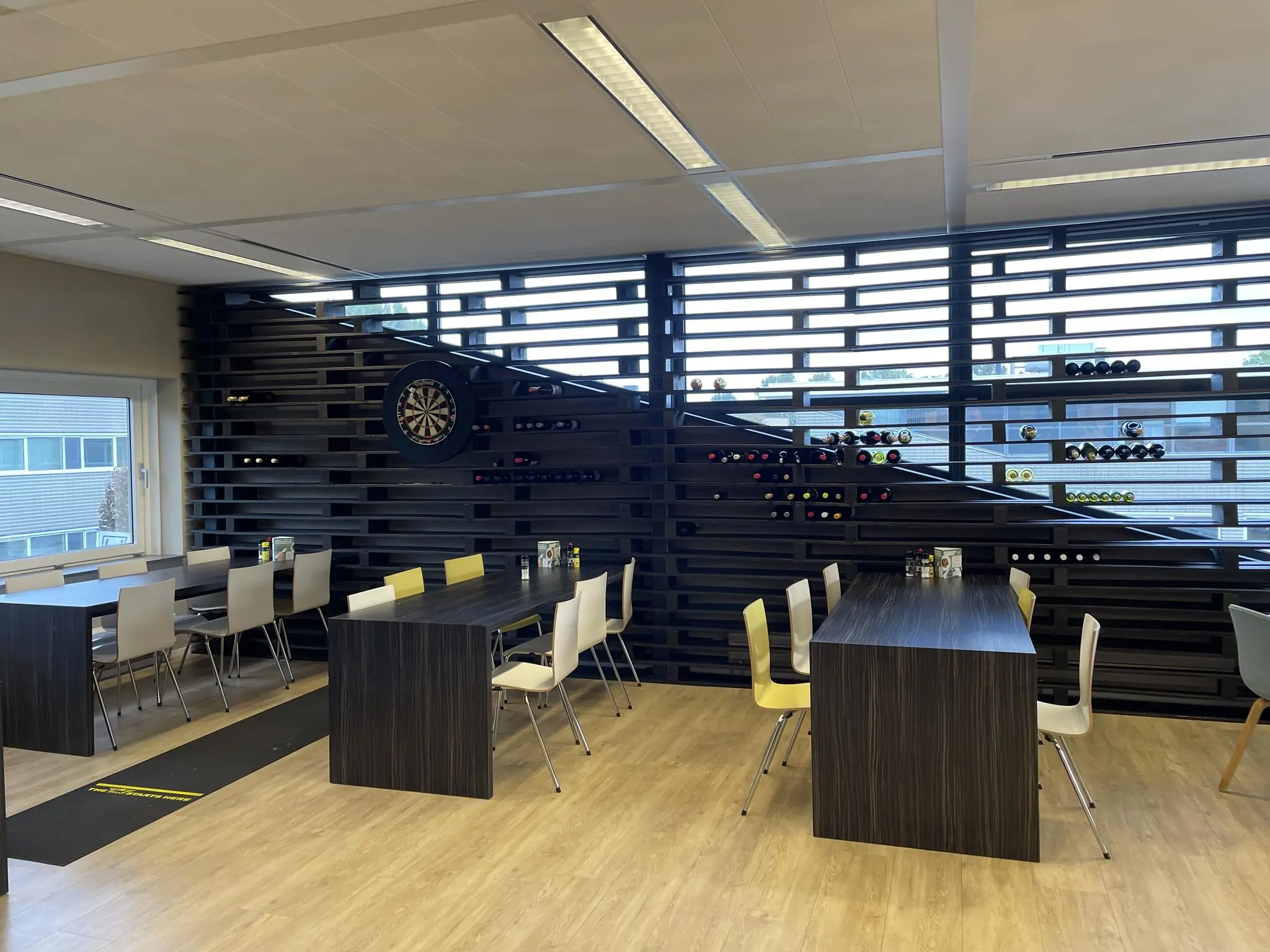 Moderne bedrijfskantine aan de Jan Leentvaarlaan met houten tafels, kleurrijke stoelen, een dartbord en een wand met wijnflessen.